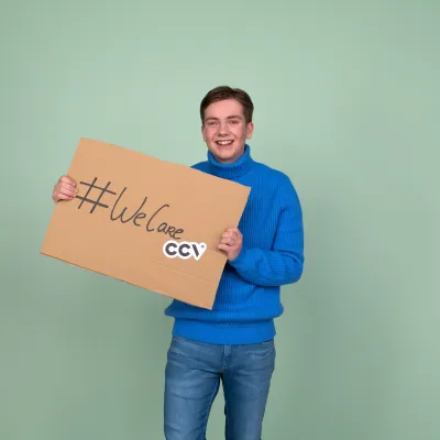 Persoon voor een groene achtergrond met een kartonnenbord. Op dit bord staat "#WeCare CCV". Persoon voor een groene achtergrond met een kartonnenbord. Op dit bord staat "#WeCare CCV".